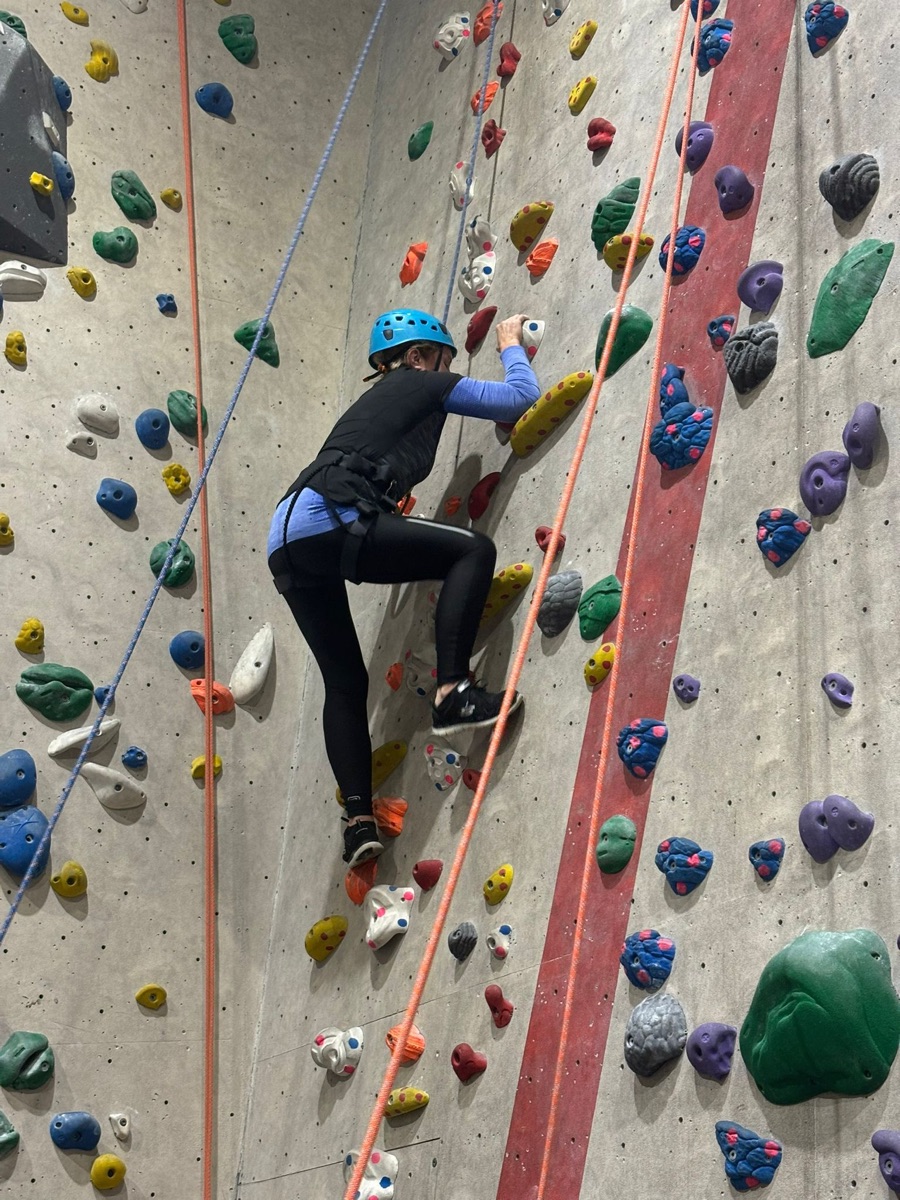 A female club member in a blue helmet and black harness part-way up an indoor climbing wall, holding a white hold in her right hand and standing on an orange hold with her left foot. Belay ropes run down the wall beside her.